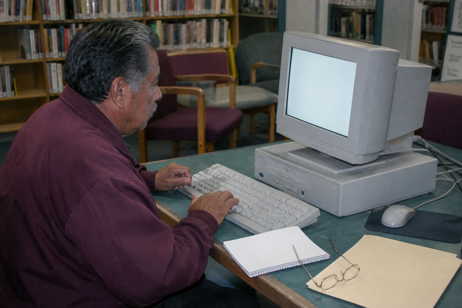 An older man using a public library computer with a notebook and folder beside him.